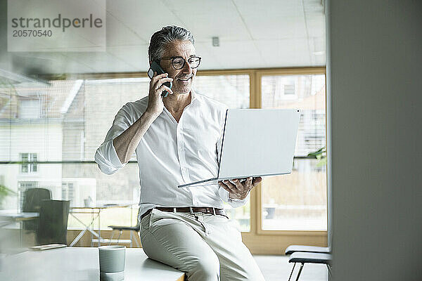 Confident businessman talking on mobile phone and using laptop in office