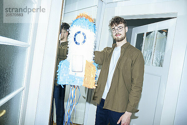 Handsome young man standing with rocketship shaped pinata at home