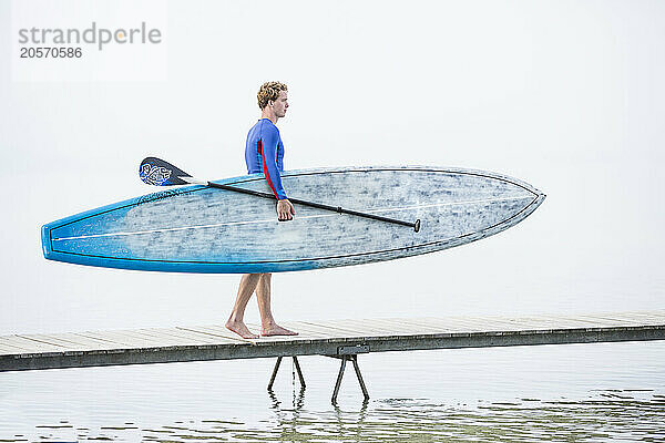 Young man carrying paddleboard and oar on jetty over lake in foggy weather