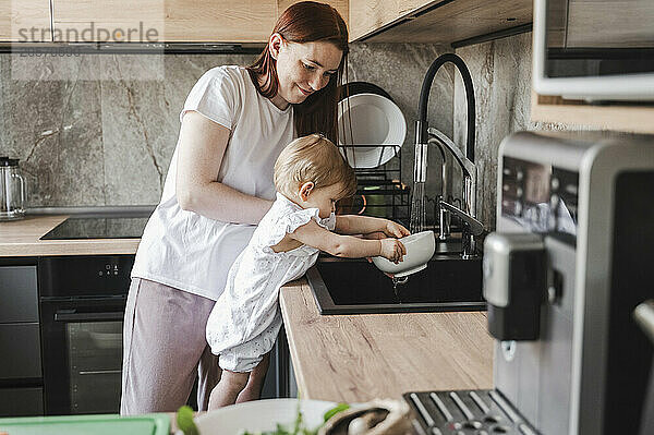 Mother and daughter washing bowl in kitchen sink together at home