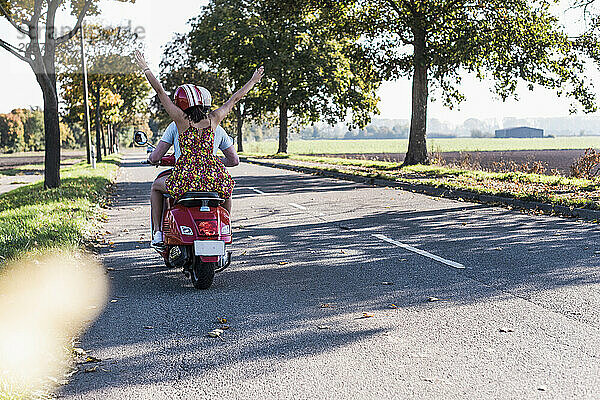 Carefree young woman with arms raised sitting on motor scooter with boyfriend at country road