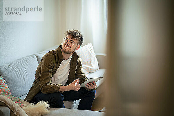 Smiling young man using tablet PC sitting on sofa at home