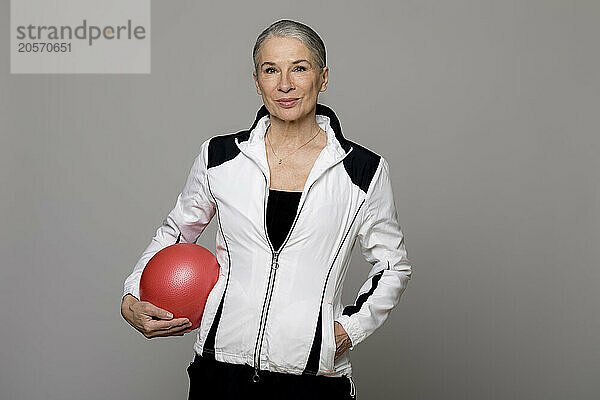 Smiling active senior woman with red ball standing against gray background