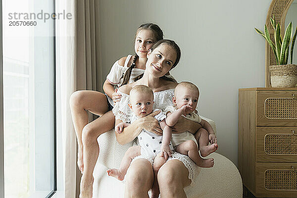 Smiling mother sitting on chair with sons and daughter at home