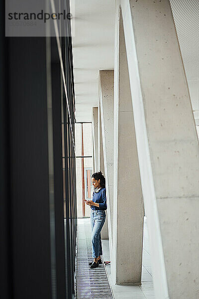 Young businesswoman using smart phone standing by columns in office