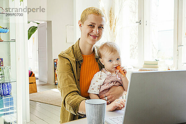 Smiling freelancer mother with daughter eating carrot at home