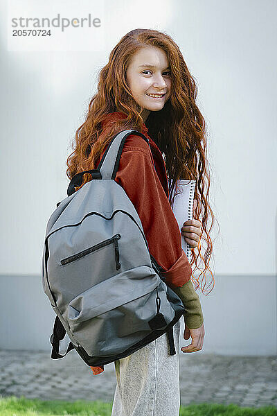 Smiling long hair redhead girl with backpack standing in front of wall