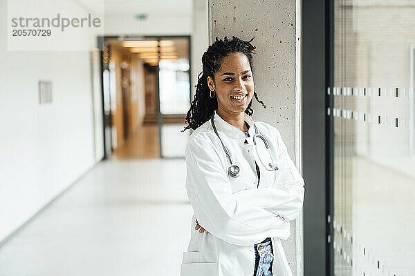 Happy young female doctor with arms crossed leaning on column in hospital corridor