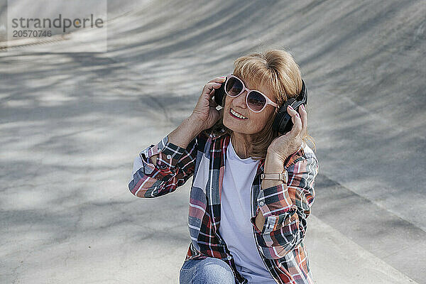 Smiling senior woman wearing wireless headphones and listening to music in skateboard park