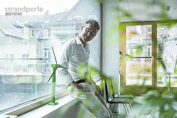 Smiling businessman leaning on window sill and looking at wind turbine model in office