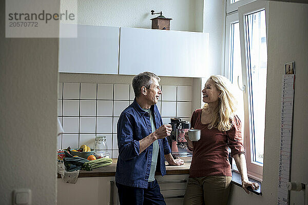 Smiling senior couple with coffee cups talking in kitchen at home