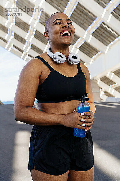 Happy muscular woman holding energy drink bottle on road