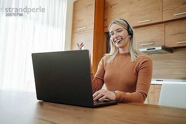 Happy young businesswoman talking through headset sitting with laptop at table in kitchen