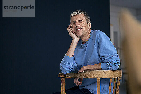 Smiling thoughtful retired man sitting on wooden chair at home