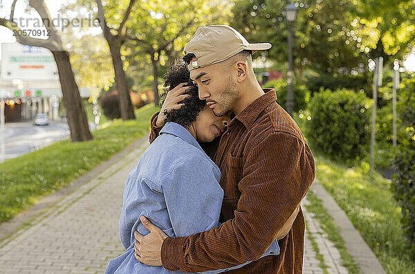 Young man wearing cap embracing girlfriend and standing at park