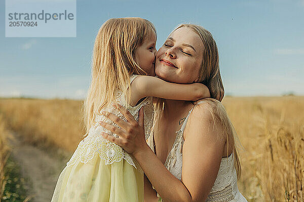 Blond hair girl kissing sister in wheat field under sky