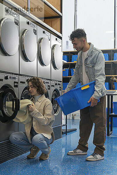 Smiling young man carrying blue laundry basket standing near girlfriend removing sheet from washing machine