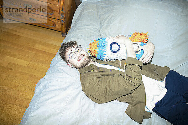 Young handsome man hugging rocketship shaped pinata lying on bed at home