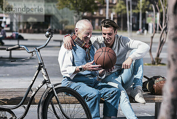 Smiling young man with grandfather holding basketball and sitting on seat at street