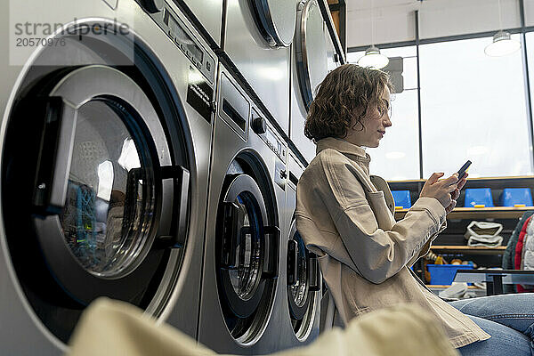 Young woman sitting in front of washing machine using mobile phone at laundromat