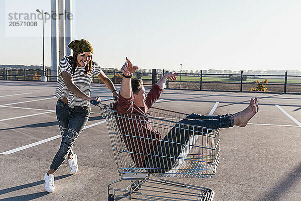 Young woman pushing boyfriend sitting in shopping cart at parking lot