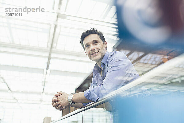 Smiling mature businessman with hands clasped leaning on railing at airport terminal