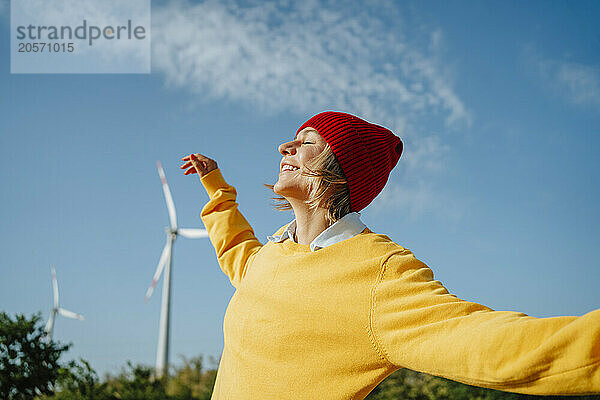 Smiling woman with orange knit hat and arms outstretched standing at wind farm