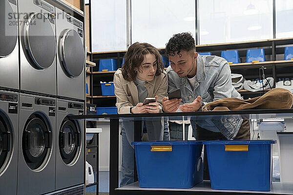 Young man and woman using smart phones leaning on table at laundromat