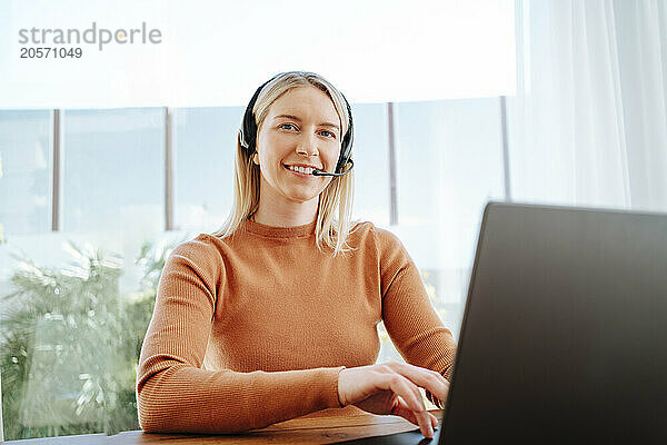 Smiling young customer service representative with headset and laptop at table working from home