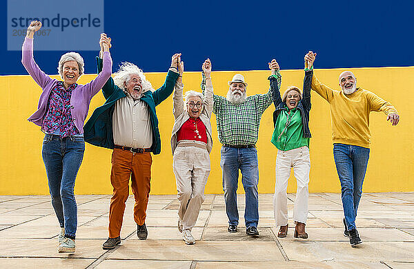 Group of elderly cheerful friends holding hands and running on footpath