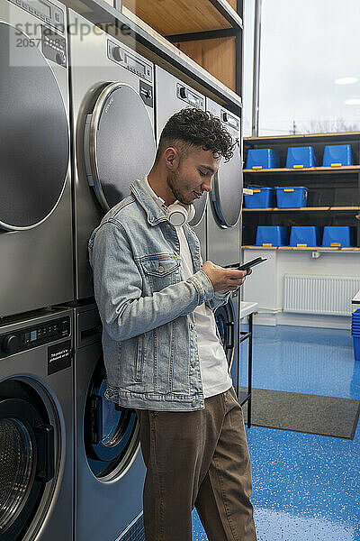 Young man leaning on washing machine using smart phone in laundromat