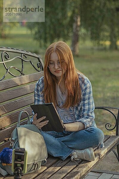 Redhead teenage girl using tablet PC sitting cross-legged on bench and vlogging through smart phone at park