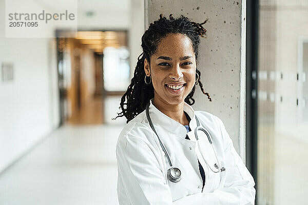 Happy young female doctor wearing lab coat leaning on column in hospital corridor