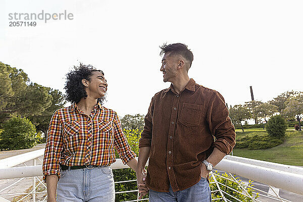 Happy boyfriend and girlfriend holding hands and standing near railing at park