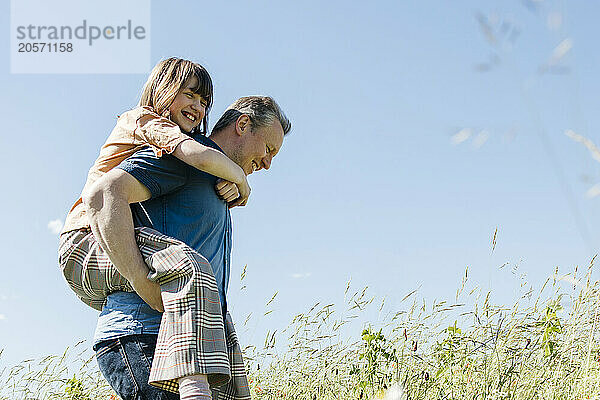 Happy father piggybacking daughter in meadow under blue sky