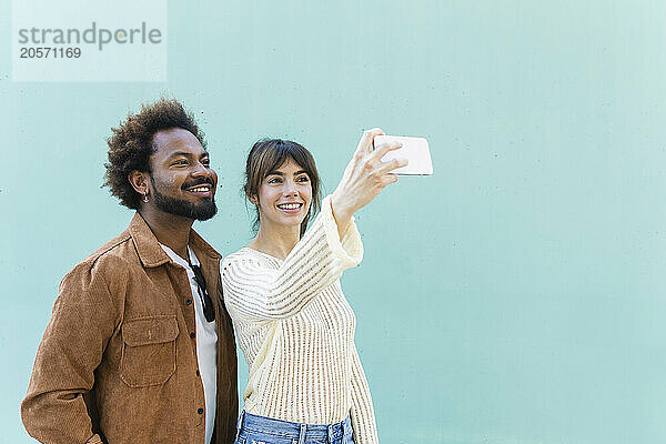 Happy woman taking selfie with boyfriend from smart phone in front of blue wall