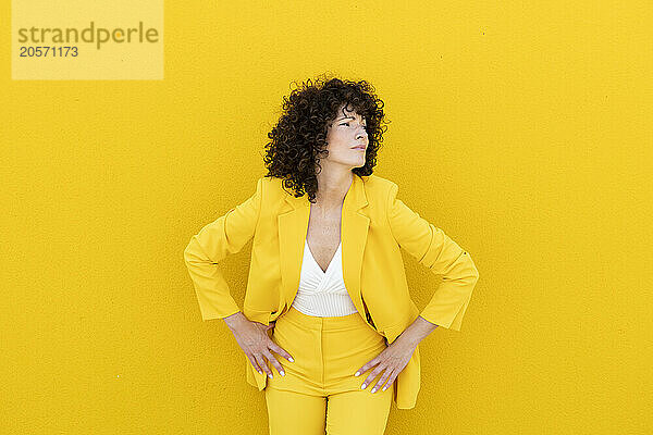 Confident woman with arms akimbo wearing blazer standing in front of yellow wall