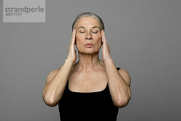 Senior woman with eyes closed touching head against gray background