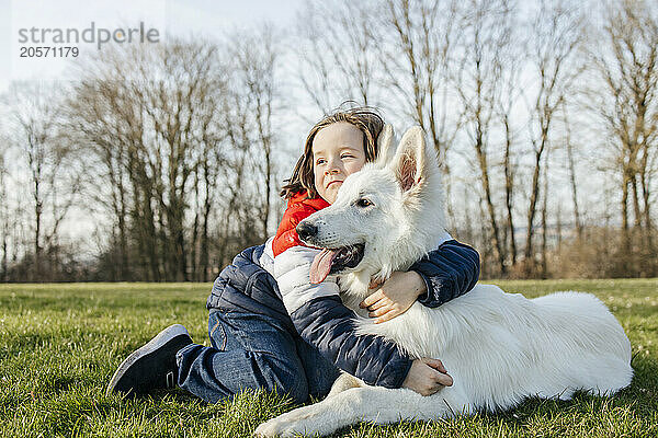 Boy with arm around White Swiss Shepherd Dog on grass in meadow