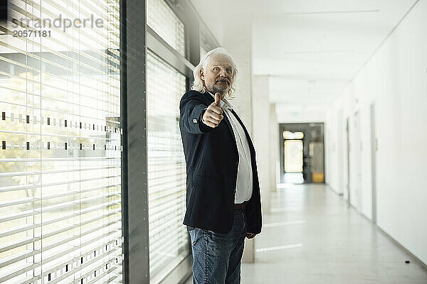 Confident senior businessman showing thumbs up standing in office corridor