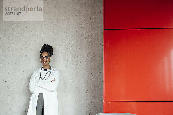 Happy young female doctor with arms crossed leaning on wall at hospital