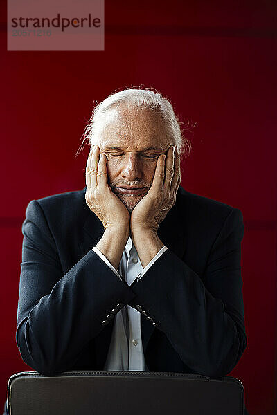 Tired senior businessman with head in hands sitting in front of red wall