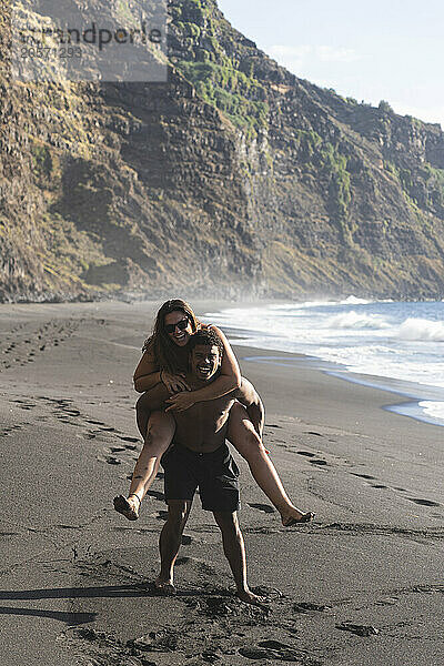 Cheerful young man piggybacking girlfriend on beach in Tenerife of Canary island