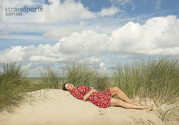 Mature woman relaxing on sand near plants under cloudy sky