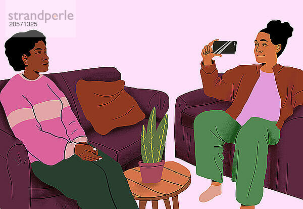 Woman photographing friend sitting on couch in front of pink background