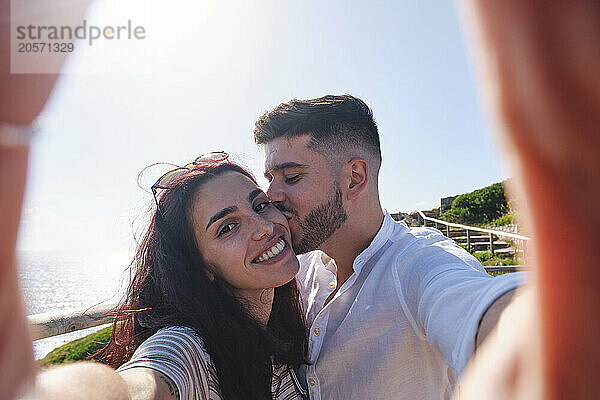 Man kissing girlfriend taking selfie at sunny day