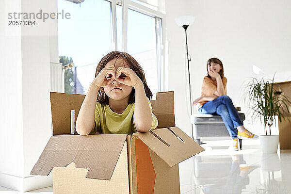 Boy in cardboard box looking through his hands with sister sitting at new house