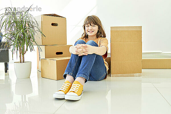 Smiling girl sitting next to boxes and potted plant on floor in new home