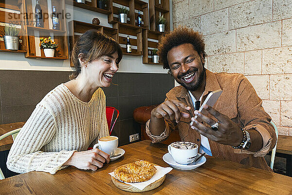 Cheerful man sharing smart phone to girlfriend sitting in cafe