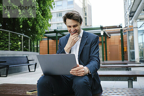 Smiling businessman with hand on chin using laptop in office park